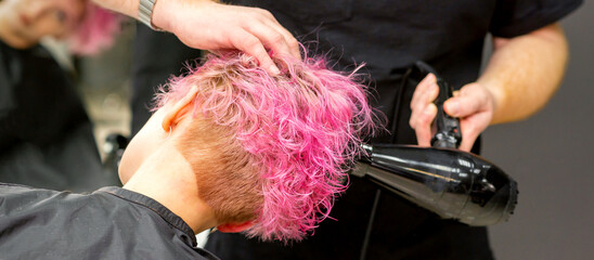 Drying short pink bob hairstyle of a young caucasian woman with a black hair dryer with the brush...
