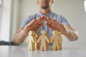 Young man holding hands above small wooden toy human figures placed on white desk as metaphor for human rights protection and safe community of people. Close up, closeup. Society, care, safety concept - obrazy, fototapety, plakaty