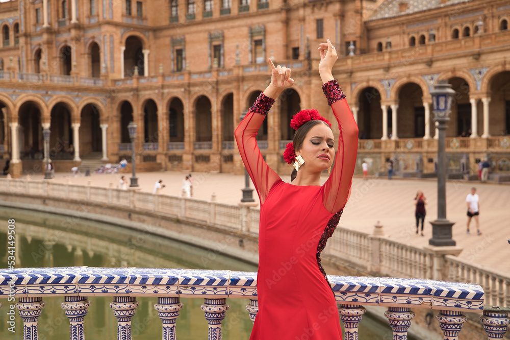 Wall mural Young teenage woman in red dance suit with red carnations in her hair doing flamenco dance poses. Flamenco concept, dance, art, typical Spanish dance.