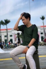 Fototapeta premium a man carrying a backpack, wearing an army green t-shirt and beige pants suffers from a headache or migraine outdoors while traveling.