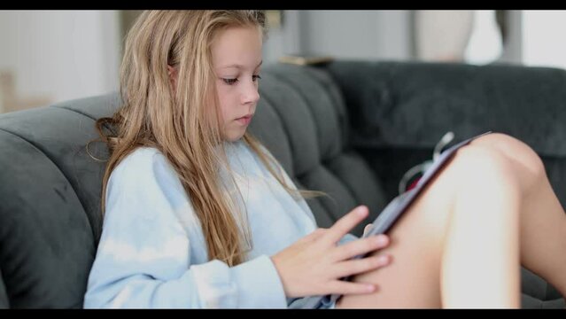 Young girl sitting at home on settee and using a tablet touch screen computer during home schooling due to closure of schools and pre-schools by coronavirus