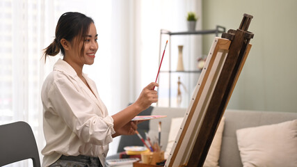 Satisfied young woman painting with watercolor on canvas in bright living room. Art, creative hobby and leisure activity concept