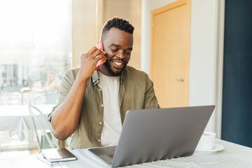Young African man talking on a cell phone while working on a computer in a coffee shop.