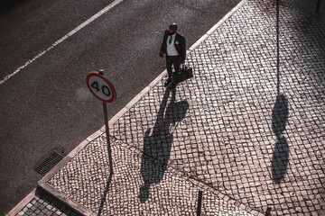 View from above of an elegant bald bearded black man entrepreneur standing on the pavement next to the road and a traffic sign casting long shadows on the asphalt and waiting for his taxi to come