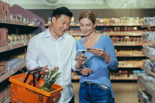 Family shopping. Young beautiful interracial couple shopping in a supermarket. Happy Asian woman and man holding credit card and phone, paying cashless, making online order.