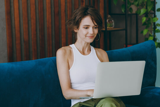 Young IT woman wears white tank shirt hold use work on laptop pc computer sit on blue sofa couch stay at home hotel flat rest relax spend free spare time in living room indoors People lounge concept.