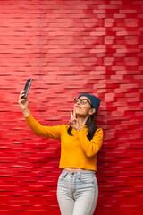 Hispanic young woman wearing an urban outfit while taking a photo of herself with her mobile phone making the victory gesture with her hand with a red wall in the background