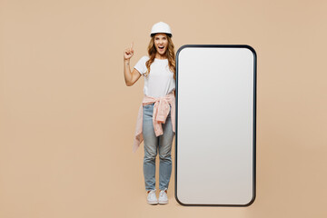 Full body young employee handyman woman in white t-shirt helmet big blank screen area mobile cell phone point finger up isolated on plain beige background Instruments accessories for renovation room.