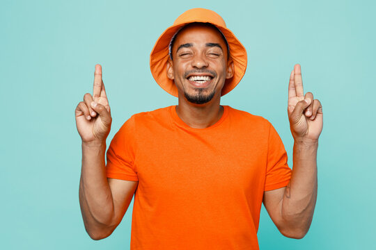 Young man of African American ethnicity wear orange t-shirt hat wait for special moment, keep fingers crossed eyes closed isolated on plain pastel light blue cyan background. People lifestyle concept.
