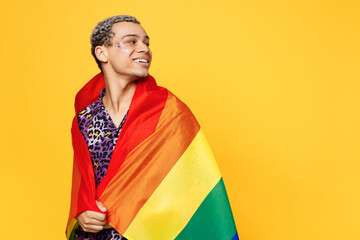 Side view young gay man wearing purple print shirt wrapped in colorful striped rainbow flag look aside on workspace area isolated on bright plain yellow color background Lifestyle lgbtq pride concept