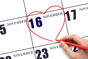 A woman's hand drawing a red heart shape on the calendar date of 16 November. Heart as a symbol of...