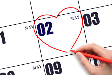 A woman's hand drawing a red heart shape on the calendar date of 2 May. Heart as a symbol of love.