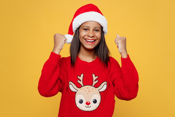 Merry fun little kid teen girl 13-14 years old wear red xmas sweater Santa hat posing do winner gesture celebrate clench fists isolated on plain yellow background. Happy New Year 2023 holiday concept.