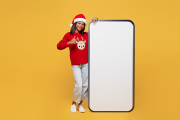 Merry little kid teen girl 13-14 years old wear red xmas sweater Santa hat posing big blank screen mobile cell phone with area isolated on plain yellow background. Happy New Year 2023 holiday concept.