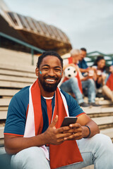 Happy black soccer fan using smart phone on stadium and looking at camera.