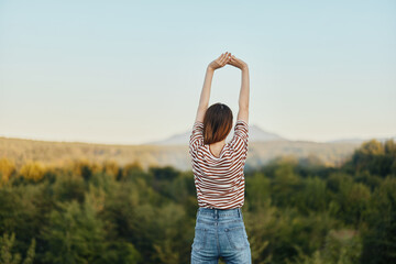 A young woman stands with her back to the camera with her hands up in a T-shirt and jeans in nature...