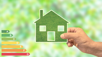 Hand holds an eco green house, with energy efficiency rating. Blurred leaves in the background