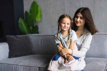 Close up happy attractive mother hugging cute preschool daughter. Smiling young mom closed eyes embracing little girl enjoying free time together sitting on couch at home.