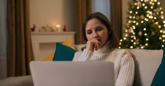 Attractive focused cheerful girl watching video comedy movie on laptop relaxing at home n couch by Christmas tree modern loft industrial interior inside.