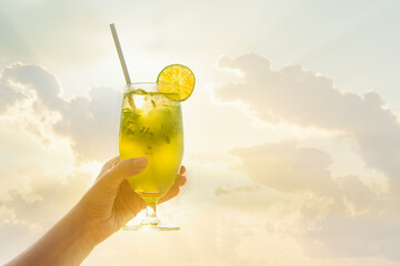 Hand holding glass of mojito on background of sun and cloud sky.