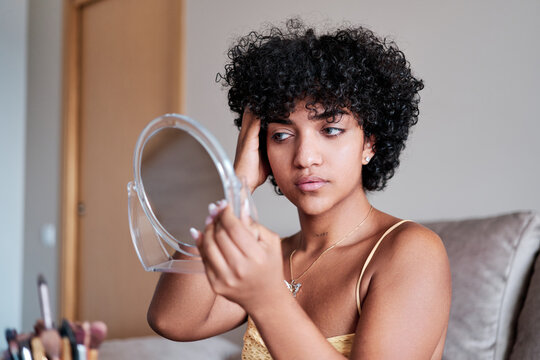 Transgender woman looking in a small mirror and combing her hair while preparing at home.