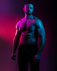 Sportive man bodybuilder is posing in the colorful neon light with naked muscular torso showing chest, abdominal muscles in neon studio light.