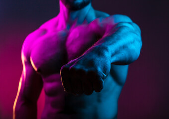 Sportive man bodybuilder shows fist in the colorful neon light with naked muscular torso, abdominal muscles in neon studio light focus on the fist