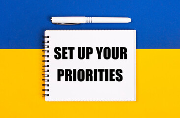 Fototapeta premium A white notebook with the text SET UP YOUR PRIORITIES and a white pen on a blue and yellow background.