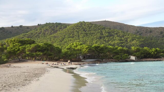 The waves splashing on beach coastline of Cala Agulla (Cala Ratjada) on Mallorca in the mediterranean sea. Deep blue water and a green pine forest in the background. People bathing,  4k.