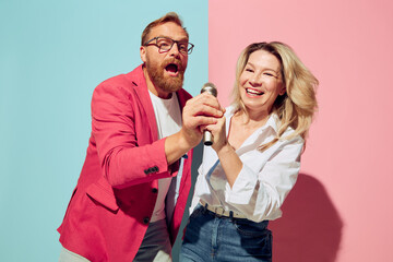 Fototapeta At karaoke. Expressive young people, man and woman, husband and wife spending time together isolated over blue and pink background. obraz