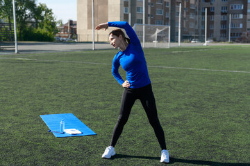 Women and sport. Girl in sportswear does exercises: bends and stretches on the grass at the stadium...