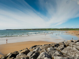 Yellow sand and blue ocean and sky. People swim in the background and surf on board. Lahinch beach in county Clare, Ireland. Warm sunny day. Irish landscape. - obrazy, fototapety, plakaty