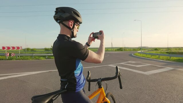 Young caucasian bicyclist in activewear using modern smartphone to take photo of incredible beautiful sunset. Concept of cycling, triathlon and modern technology.