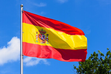The national flag of Spain consists of red, yellow and red horizontal stripes with the coat of arms in the middle on a background of blue sky in the afternoon in summer