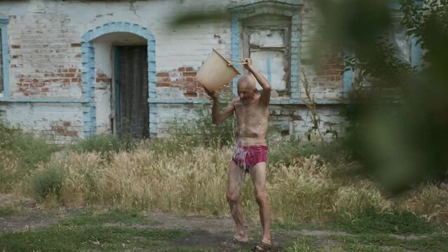 Skinny elderly man pours cold water on himself out of bucket. Pensioner hardens body and health standing in yard against old brick house slow motion
