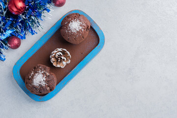 Chocolate cupcakes and a pine cone on a platter next to a garland and baubles on marble background