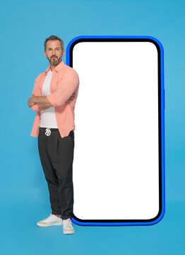 Posing with arms crossed in front big smartphone with white screen in blue case middle aged grey haired man wearing peach shirt and jogger pants isolated on blue background. Mobile app advertising