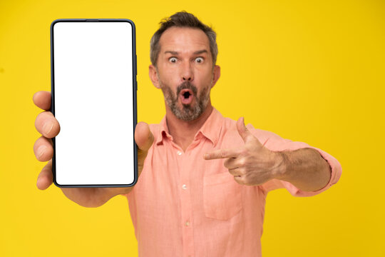 Excited, shocked of a new mobile app middle aged grey haired man pointing at smartphone with white screen in hand wearing peach shirt isolated on yellow. Mobile app advertisement
