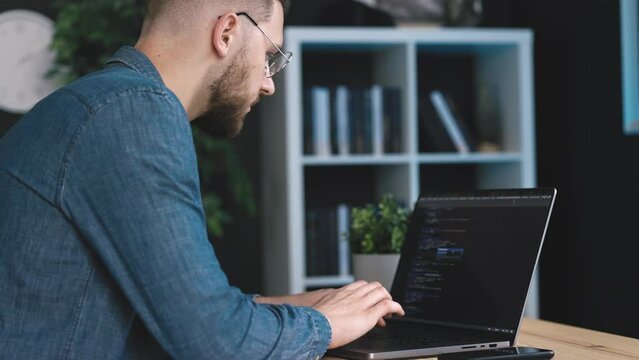 Focused bearded man in denim shirt writing code on laptop, young programmer coding