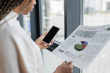 Cropped view of african american businesswoman holding mobile phone and paper with charts in office.