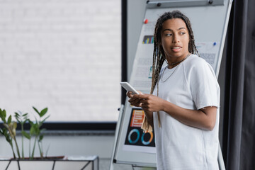 African american businesswoman in t-shirt holding smartphone near blurred flip chart in office.