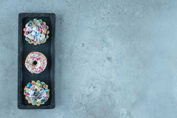 Candy-topped cupcakes and donuts on a black tray on marble background