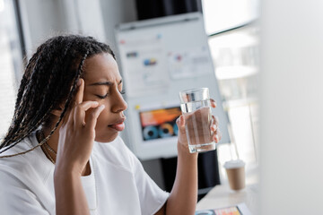 African american businesswoman holding glass of water while suffering from headache in office.