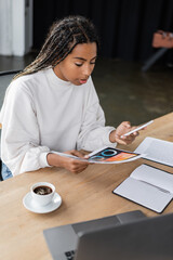 African american businesswoman using smartphone and holding paper with charts near coffee in office.