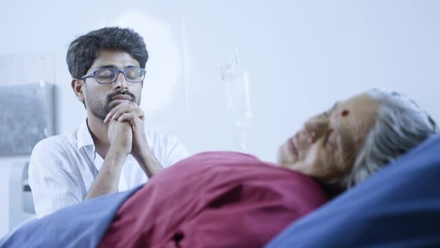 Worried indian son praying god for sick mother for speed health recovery at hospital- concept of healthcare, unconditional bonding and treatment.