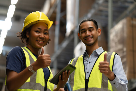 Black woman with yellow helmet and man warehouse worker show thumb up together for successful job with happy face. Team spirit working together in storehouse to complete the balance inventory