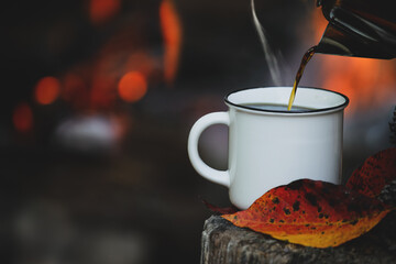 Kettle pouring hot steaming coffee into a white enamel cup  sitting on an old log with autumn leaves. Selective focus on mug with blurred foreground and background.