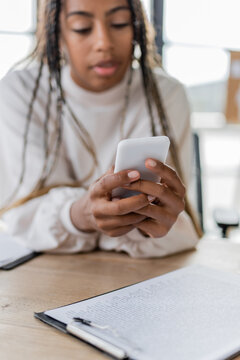 Blurred african american businesswoman using smartphone near clipboard in office.