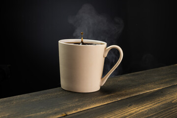 A mug of hot coffee with evaporation and steam and a small splash on a wooden table against a dark background