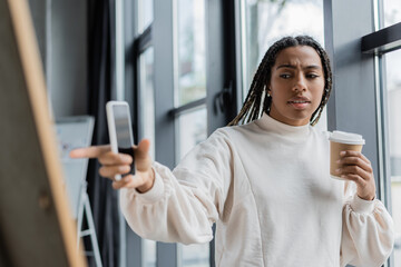 African american businesswoman holding coffee to go and smartphone while pointing at blurred board in office.